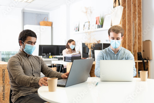 Coworkers concentrated on their laptops and wearing protective face masks. Working in the office during Coronavirus pandemic concept.