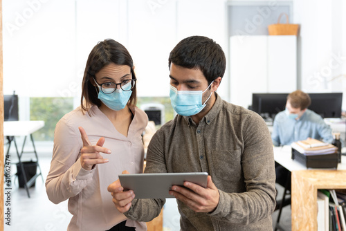 Hispanic man and woman wearing protective face masks chatting while looking a tablet. Working in the office during Coronavirus pandemic concept.