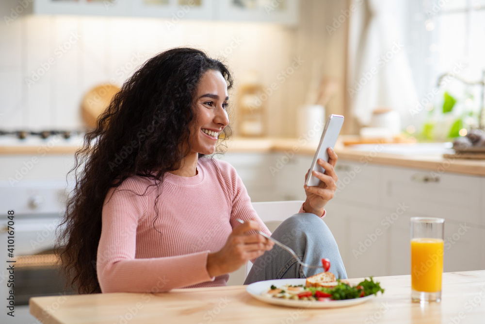 Happy Young Woman Using Smartphone In Kitchen While Having Tasty Breakfast
