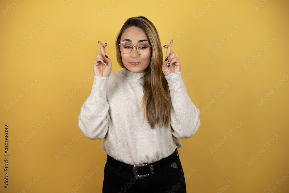Pretty woman with long hair gesturing finger crossed smiling with hope and looking side against yellow wall Pretty woman with long hair gesturing finger crossed smiling with hope and looking side against yellow wall