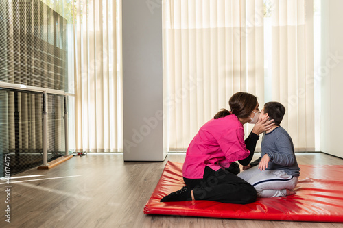 disabled child and physiotherapist give a kiss on a red mat doing exercises. pandemic mask protection