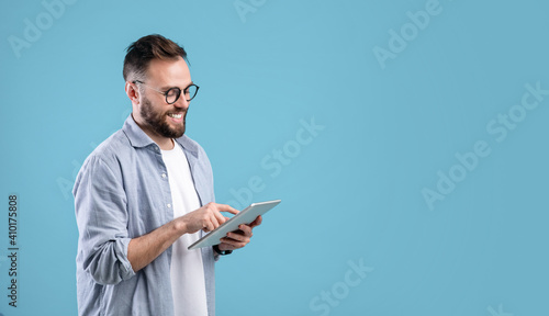 Smart millennial guy in glasses using tablet computer over blue studio background, banner design with copy space