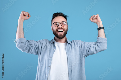 Overjoyed bearded guy in glasses making YES gesture with both hands on blue studio background, panorama