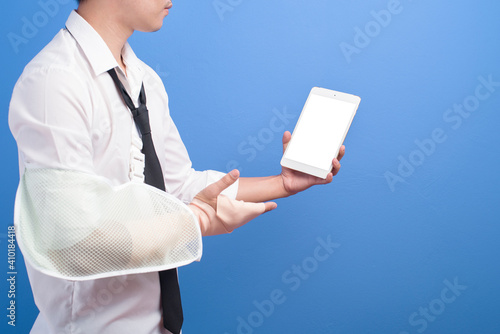 Young businessman with an injured arm in a sling using a tablet over blue background in studio, insurance and healthcare concept
