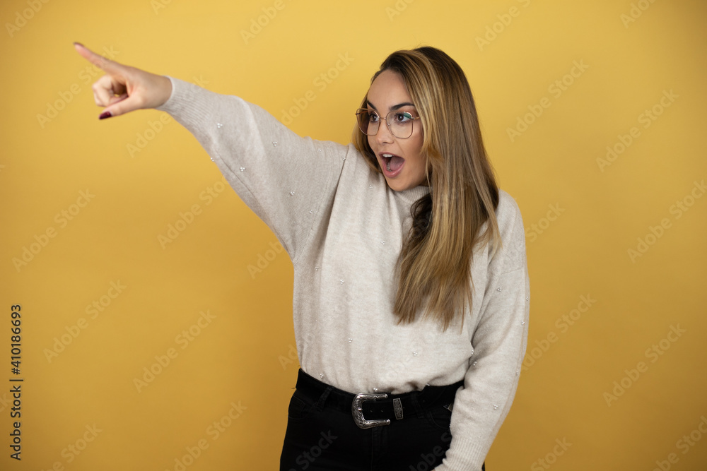 Pretty woman with long hair pointing with finger surprised ahead, open mouth amazed expression, something on the front against yellow wall Pretty woman with long hair pointing with finger surprised ahead, open mouth amazed expression, something on the front against yellow wall