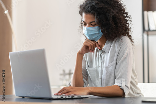 A thoughtful young African American businesswoman with curly hair wearing a protective face mask sitting at the desk and working on a laptop, back to the office after covid-19 lockdown concept