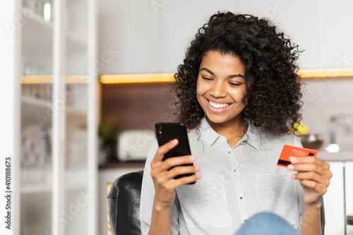 Smiling African American teenage girl with curly hair holding mobile phone, entering credit card number to make an online transaction, mixed-race woman ordering food, doing online shopping from home