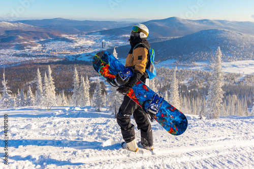 A male snowboarder holds a blue snowboard and looks up at the sky on a sunny frosty day