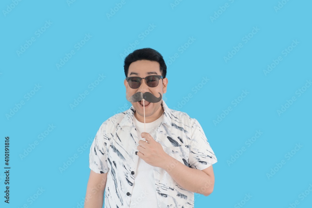 Smiling Asian man in white casual shirt holding fake mustaches stick over mouth over blue background. Smiling Asian man in white casual shirt holding fake mustaches stick over mouth over blue background.