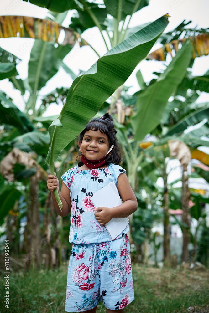 Little schoolgirl wearing face mask and covering head with banana leaf going to school Little schoolgirl wearing face mask and covering head with banana leaf going to school