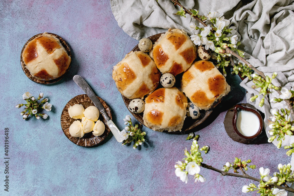 Fototapeta premium Homemade Easter traditional hot cross buns on ceramic plate with blossom cherry branches, butter, jug of milk, textile over blue texture background. Flat lay, space