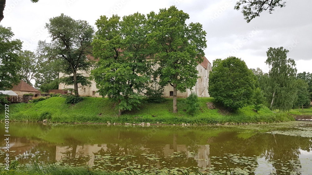 Latvian castle Jaunpils behind green trees on the shore of a small lake on July 12, 2019 Latvian castle Jaunpils behind green trees on the shore of a small lake on July 12, 2019
