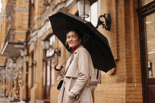 Snapshot of charming Asian girl in stylish trench coat holding black umbrella on city street