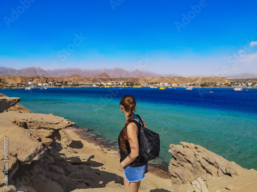 A young woman stands on a rock and looks at the opposite side of the Sharm el-Maya bay in Sharm El Sheikh (Egypt) - a view from the back. Tourist among the stones on the shores of the Red Sea