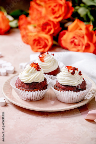 Homemade Red velvet cupcakes with whipped cream on pink ceramic plate, white napkin with ribbon, roses flowers, wooden hearts over pink texture background. Valentines day dessert.