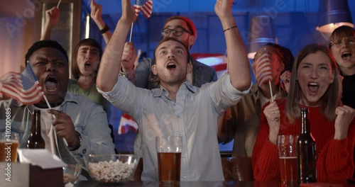 Diverse people with american flags and shouting while celebrating national team success. Excited multiethnic people rejoicing and smiling while watching sportrs game with beer in pub photo