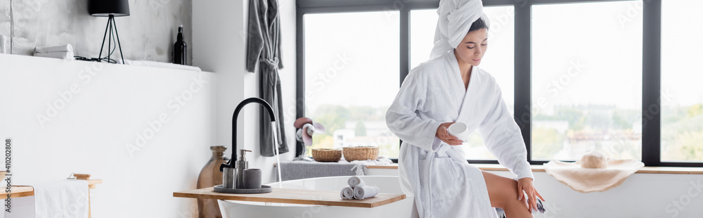 Woman in bathrobe applying cosmetic cream on leg in bathroom, banner - obrazy, fototapety, plakaty 