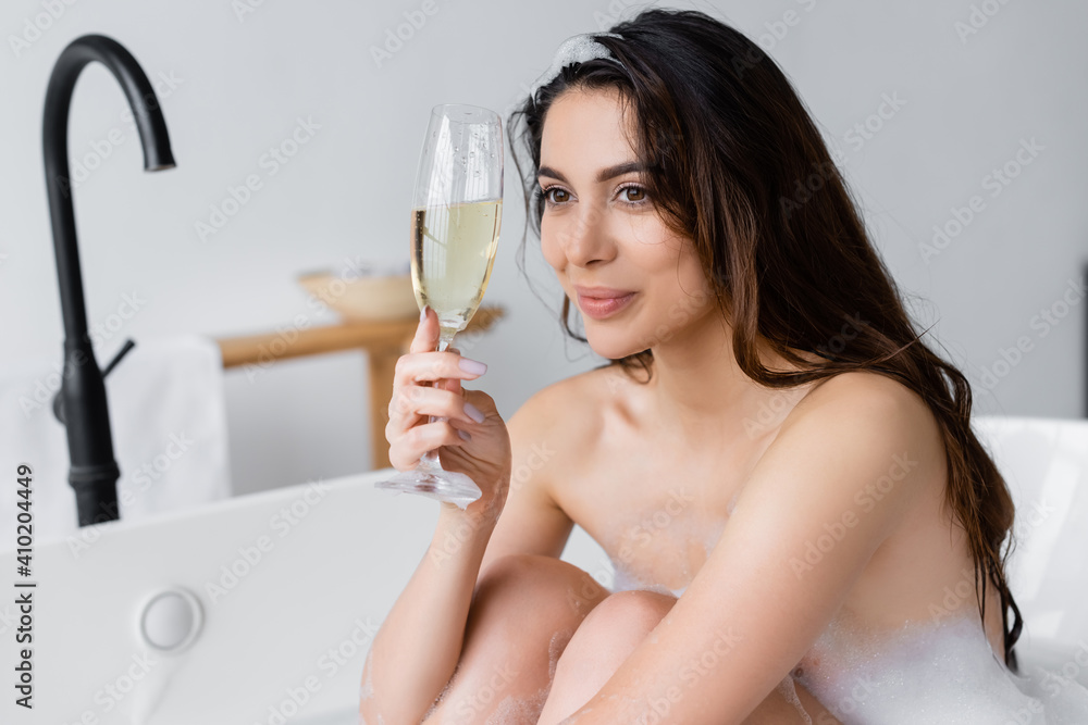 Brunette woman with foam on hair holding champagne glass in bathtub Brunette woman with foam on hair holding champagne glass in bathtub