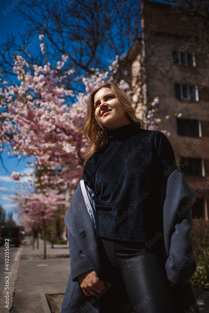 Sakura branches with flowers on a tree on the city streets. Happy woman girl in a gray palette walks along an alley with blooming sakura. Gorgeous fancy girl outdoors. Sakura tree blooming. Sakura branches with flowers on a tree on the city streets. Happy woman girl in a gray palette walks along an alley with blooming sakura. Gorgeous fancy girl outdoors. Sakura tree blooming.