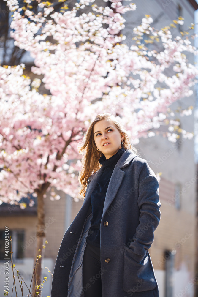 Sakura branches with flowers on a tree on the city streets. Happy woman girl in a gray palette walks along an alley with blooming sakura. Gorgeous fancy girl outdoors. Sakura tree blooming.