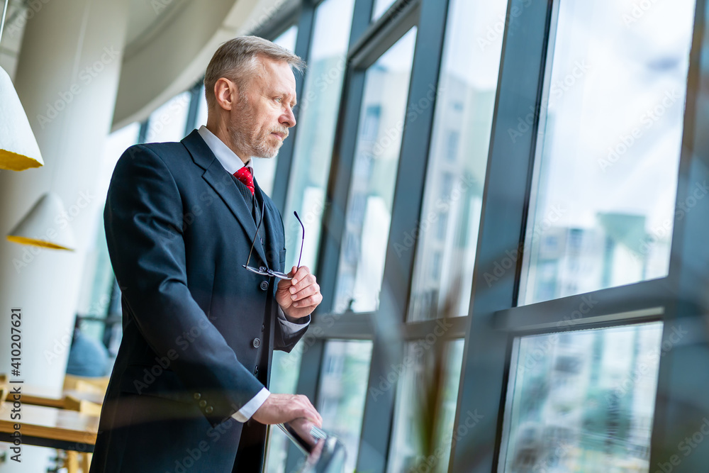 Elegant senior man in dark suit and red tie is looking to the panoramic window. View from below. Elegant senior man in dark suit and red tie is looking to the panoramic window. View from below.