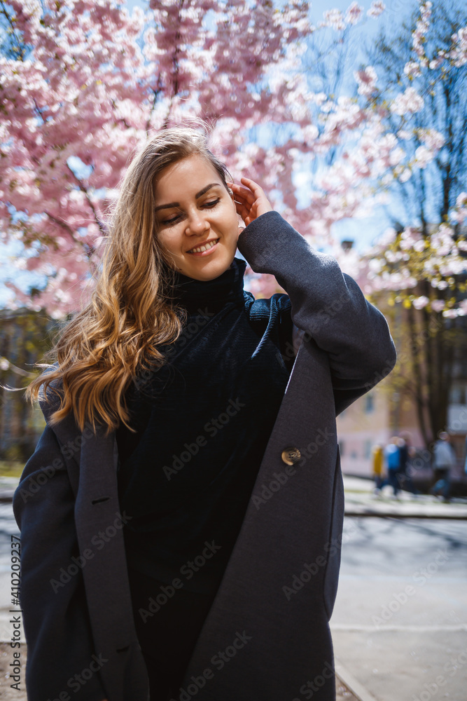 A young slender female model with long wavy hair and, dressed in a gray coat, sneakers, stands on the street near a flowering shrub with beautiful pink flowers in the background and poses. A young slender female model with long wavy hair and, dressed in a gray coat, sneakers, stands on the street near a flowering shrub with beautiful pink flowers in the background and poses.