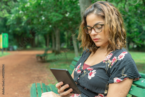 Beautiful Latin Woman with glasses and a piercing in her nose reading an E-book in a park