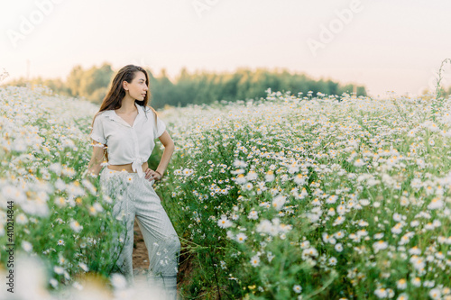 A young woman in white clothes walks along a path in the middle of a blooming chamomile - soft sunset light and the feeling of a warm summer day.