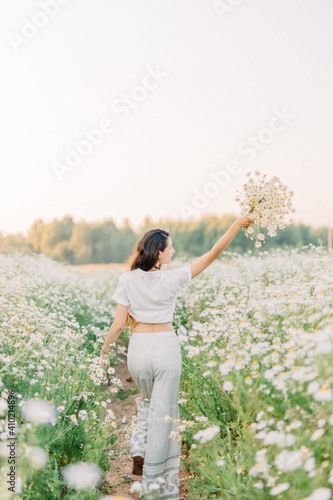 Beautiful girl with daisies bouquet, back view. Tranquil summer in countryside. Stylish young woman in white vintage dress and hat waling on field of daisies.