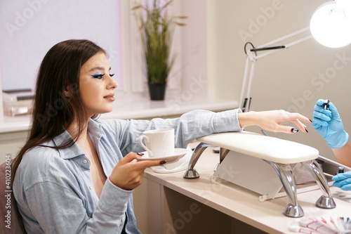 Beautiful woman getting a manicure in a beauty salon. She smiles and holds a white cup of coffee in her hand.