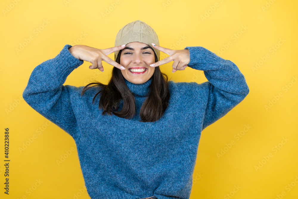 Young beautiful woman wearing blue casual sweater and wool hat Doing peace symbol with fingers over face, smiling cheerful showing victory Young beautiful woman wearing blue casual sweater and wool hat Doing peace symbol with fingers over face, smiling cheerful showing victory