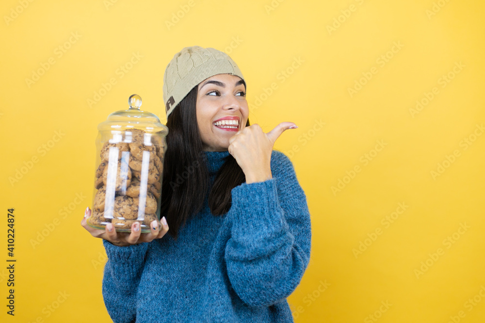Young beautiful woman wearing blue casual sweater and wool hat holding chocolate chips cookies jar smiling and pointing with hand and thumb to the side