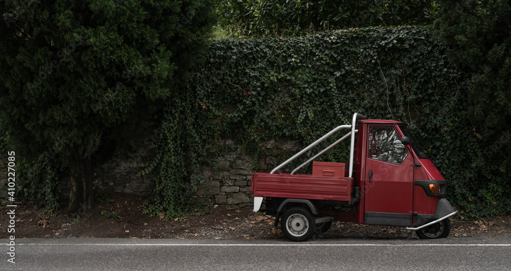 Red, little retro car or delivery truck on the road among green wall of plants. Typical red and green italian picture. in dark, dull colors.