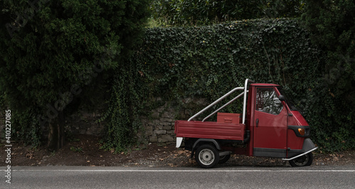 Obraz na płótnie Red, little retro car or delivery truck on the road among green wall of plants. Typical red and green italian picture. in dark, dull colors. w salonie