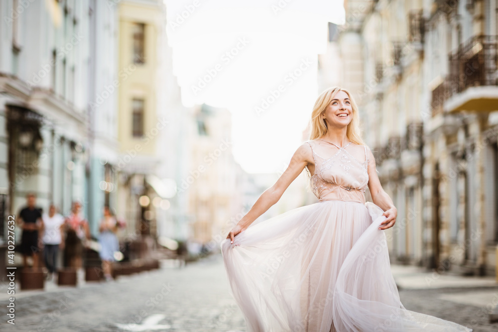 Pretty smiling young woman with long blond hair in elegant flying light dress running along the street