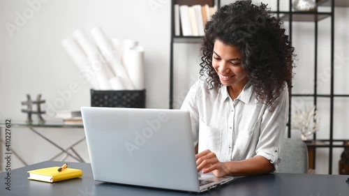 Cheerful smart african-american woman sitting at the desk and typing on the laptop, a female entrepreneur with a computer in the office satisfied with a result of work, answers emails with a smile