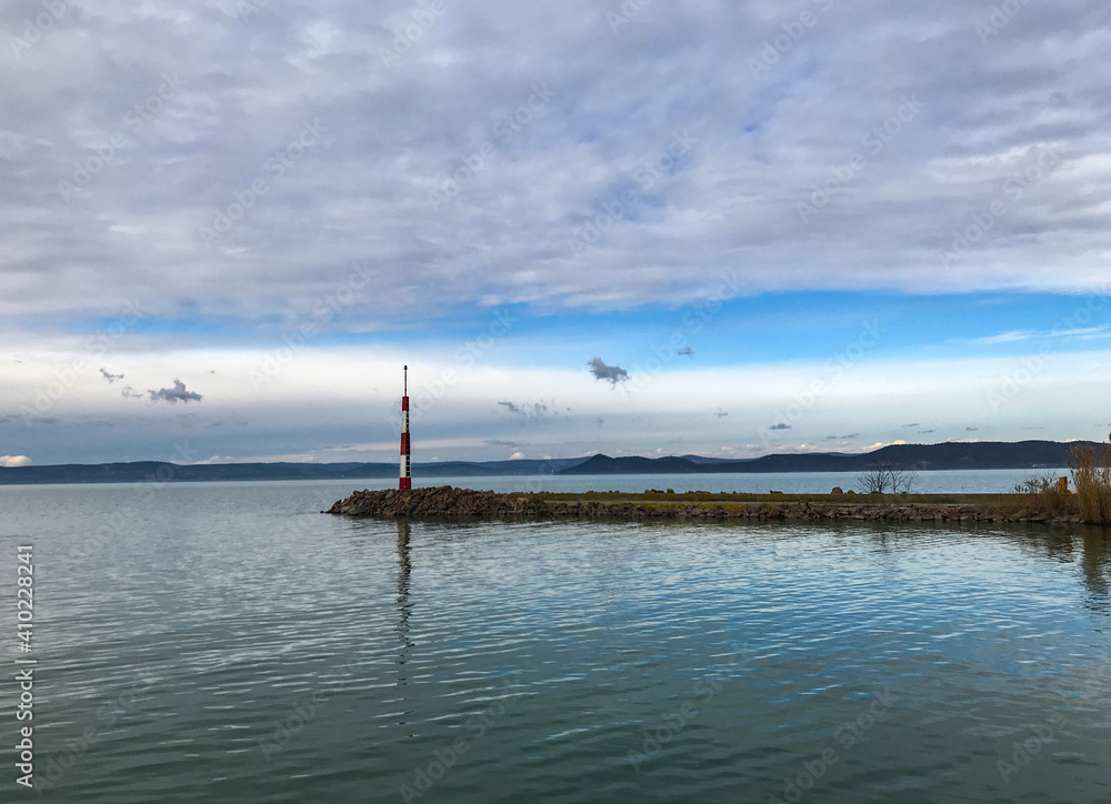 Lake pier with a lonely mooring pole with blue cloudy sky and landmarks in background. Autumn scene at lake shore