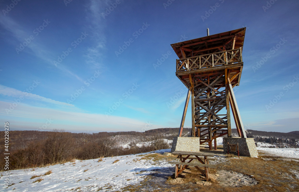 Lonely tall wooden lookout tower stands on a snowy winter field, with a wooden bench and desk.  View from below, mountains and blue sky in background. Winter landscape in a sunny day.