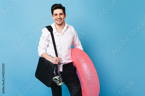 Handsome guy in white shirt is holding laptop bag. Man in glasses posing with inflatable circle and diving mask on blue background