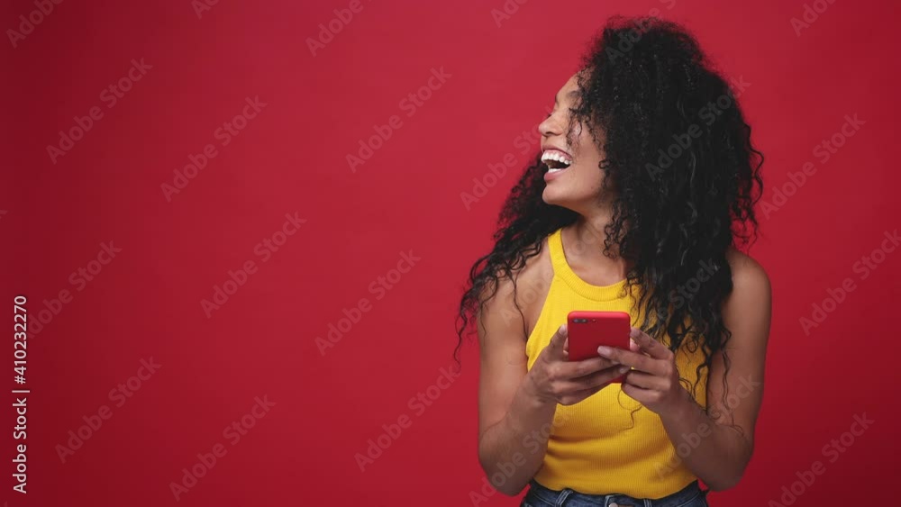 Cheerful excited african american young woman in basic yellow tank top posing isolated on red background studio. People lifestyle concept. Using mobile cell phone typing browsing looking aside say wow Cheerful excited african american young woman in basic yellow tank top posing isolated on red background studio. People lifestyle concept. Using mobile cell phone typing browsing looking aside say wow