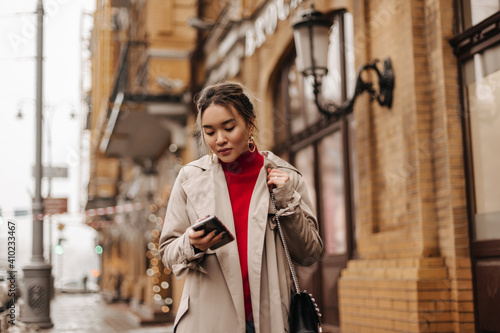 Stylish Asian lady in beige coat, red top and cross-body bag walks around city, holding smartphone