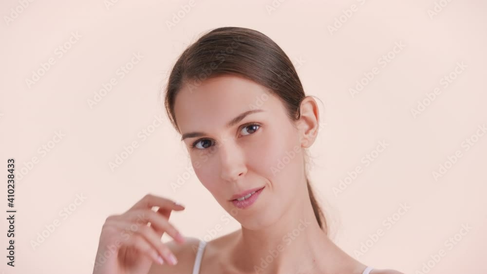 Close-up beauty portrait of young brunette woman with perfect skin gently touches her face sliding hand along a jawline down to her chin, looks at camera and smiles | Skincare products ad concept Close-up beauty portrait of young brunette woman with perfect skin gently touches her face sliding hand along a jawline down to her chin, looks at camera and smiles | Skincare products ad concept