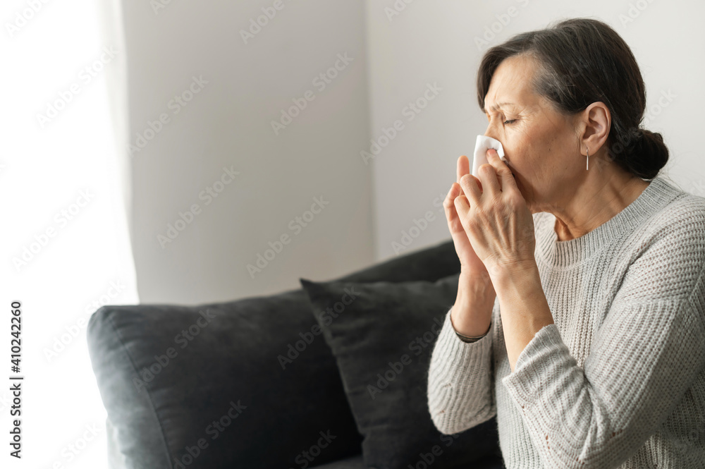 Sick senior woman has cold and flu, sneezes and has runny nose, wipes her nose with a paper tissue sitting on the couch at home. Viral disease concept Sick senior woman has cold and flu, sneezes and has runny nose, wipes her nose with a paper tissue sitting on the couch at home. Viral disease concept