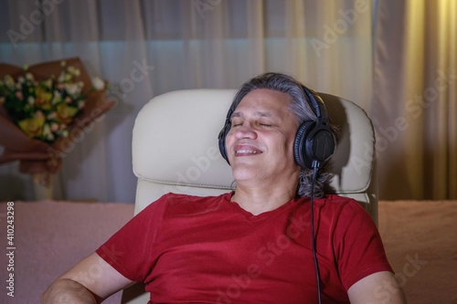 50-year-old man listens to music on headphones at home, sitting in a chair.