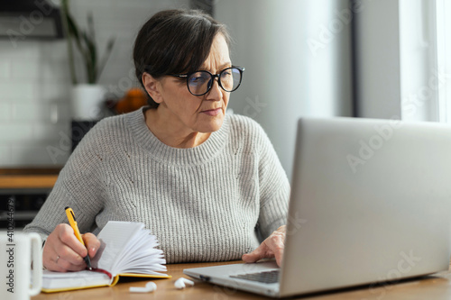 Concentrated retirement woman using a laptop sitting at the desk in the kitchen for remote work, studying online, watching a webinar and takes a notes. A senior business lady with computer indoor