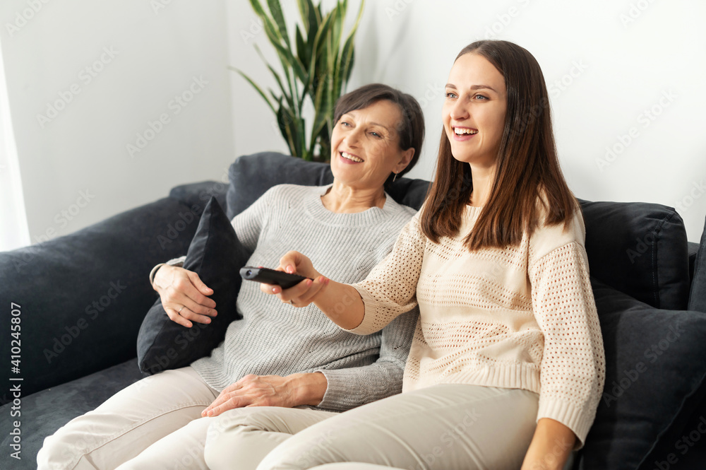 A senior mother and an adult daughter relaxing on sofa at home, watching TV, switching channels with remote control, smiling and enjoying time together A senior mother and an adult daughter relaxing on sofa at home, watching TV, switching channels with remote control, smiling and enjoying time together