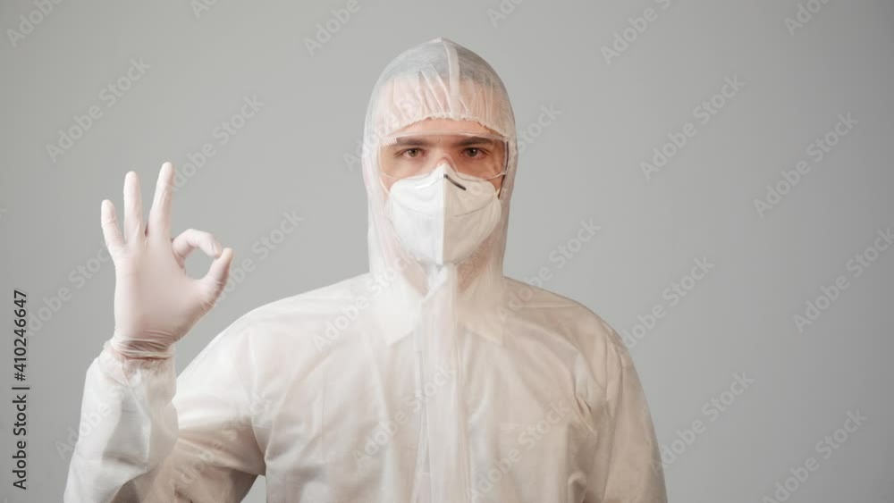 A doctor in a medical suit looks at the camera and shows a hand gesture of ok. The concept of good news from a doctor. A doctor in a medical suit looks at the camera and shows a hand gesture of ok. The concept of good news from a doctor.