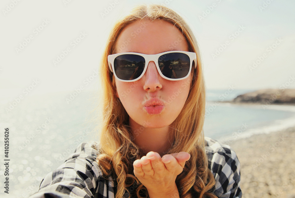 Portrait close up of happy young woman blowing her lips sending sweet air kiss stretching hand for taking selfie on a sea background Portrait close up of happy young woman blowing her lips sending sweet air kiss stretching hand for taking selfie on a sea background