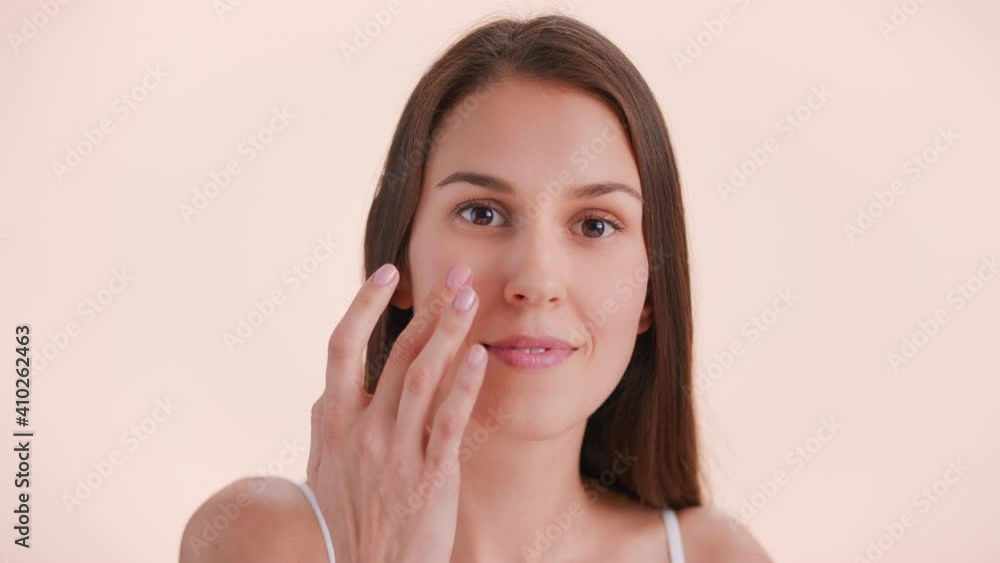 Close-up beauty portrait of young brunette woman with long hair and perfect skin applies cream on her face and smiles | Smear of moisturizing cream on woman's face | Skincare product promotion Close-up beauty portrait of young brunette woman with long hair and perfect skin applies cream on her face and smiles | Smear of moisturizing cream on woman's face | Skincare product promotion