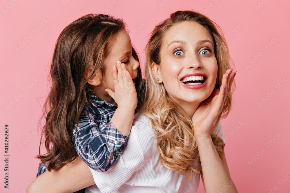 Mom and daughter gossip on pink background. Blue-eyed curly woman in white T-shirt stares into camera in amazement Mom and daughter gossip on pink background. Blue-eyed curly woman in white T-shirt stares into camera in amazement